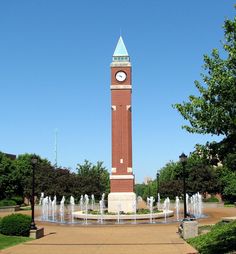 a clock tower in the middle of a park with water fountains and trees around it