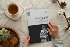 a person is reading a book next to a cup of tea and some cookies on a bed