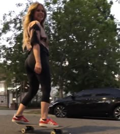 a woman riding a skateboard down a street next to a black car and trees