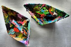 two colorful paper boats sitting on top of a metal surface with paint splatters all over them