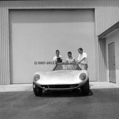 three men standing next to a car in front of a garage door with the doors open