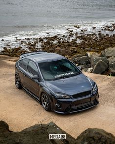 a grey car parked on top of a dirt road next to the ocean and rocks