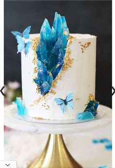 a white cake with blue butterflies on top and gold trim around the edges is sitting on a stand