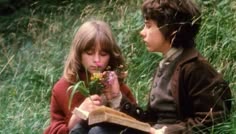 two children sitting in the grass and one is reading a book while the other holds a flower