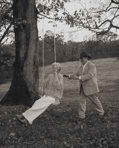 an old man and woman swinging on strings in the grass next to a large tree