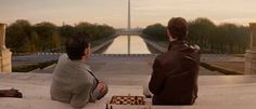 two people sitting on the ground playing chess in front of an obelisk and reflecting water