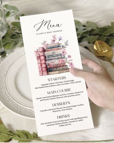 a menu for a baby shower with books on it and flowers in the background, held up by a person's hand