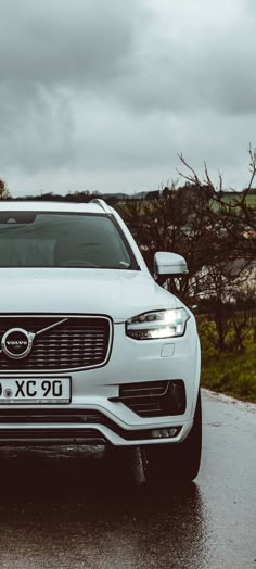 the front end of a white volvo suv parked on a wet road with trees in the background