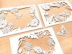 three paper cut designs with flowers and butterflies on them, sitting on a wooden surface