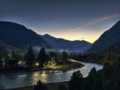 a river flowing through a lush green forest under a cloudy sky at night with mountains in the background
