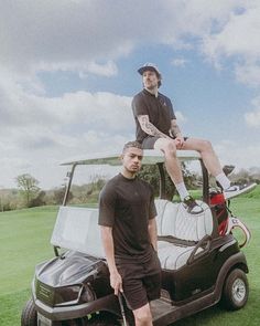 two men standing next to each other on top of a golf cart in the grass