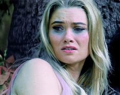 a woman with blue eyes and long blonde hair sitting in front of a tree looking at the camera