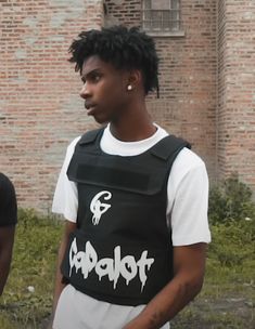 a man with dreadlocks standing in front of a brick building wearing a black vest