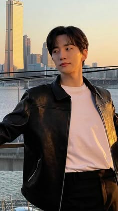 a young man standing in front of a body of water wearing a black leather jacket and white t - shirt