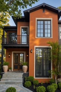 Modern two-story home with an orange exterior, large windows, and potted plants flanking the entrance, surrounded by green landscaping.