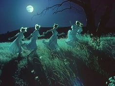 four women in white dresses are dancing on the grass at night with a full moon behind them