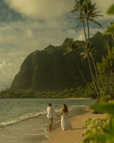 a man and woman are walking on the beach holding hands with mountains in the background