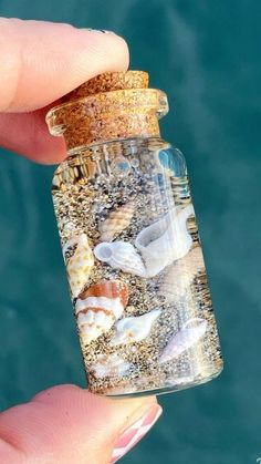 a hand holding a bottle filled with sand and sea shells on top of the ocean