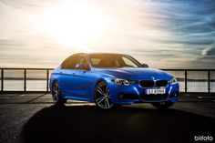a blue bmw car parked in front of the ocean with the sun shining behind it