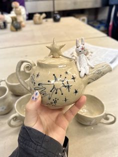a hand holding a ceramic teapot with stars on it and a cat figurine sitting on top