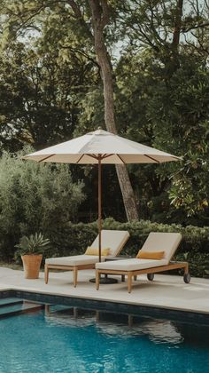 two lounge chairs under an umbrella next to a swimming pool with trees in the background