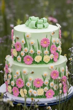 a three layer cake decorated with daisies and green ribbons on a plate in the middle of flowers