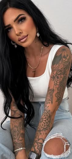 a woman with long black hair and tattoos on her arm sitting in ripped jeans looking at the camera