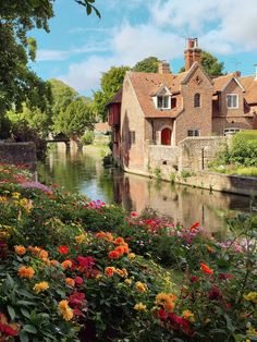 a river running through a lush green forest filled with lots of flowers next to a brick building