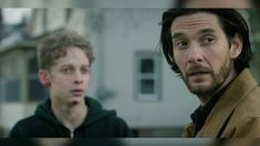 two young men standing next to each other in front of a white house and one is looking at the camera