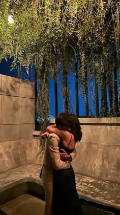 a woman is hugging another woman in front of some plants and trees at night time