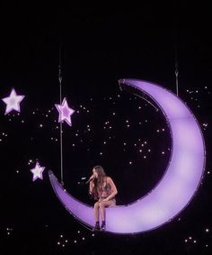 a woman sitting on top of a crescent moon with stars hanging from the ceiling behind her
