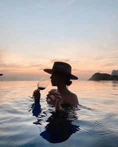 a woman holding a wine glass in her hand while sitting in the water at sunset