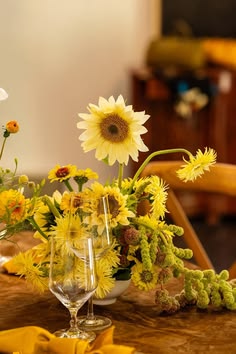a wooden table topped with two wine glasses filled with yellow and white sunflowers