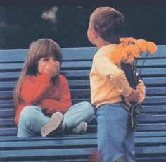 two children sitting on a blue bench and one is covering his mouth while the other holds flowers
