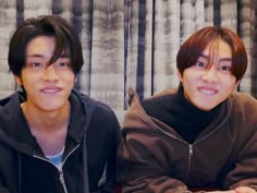 two young men sitting next to each other in front of a window with curtains behind them