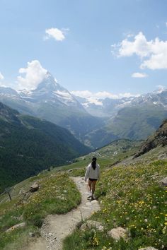 a woman walking up a trail in the mountains with flowers on both sides and snow capped peaks behind her