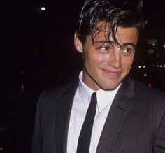 a young man in a suit and tie smiling at the camera with city lights behind him
