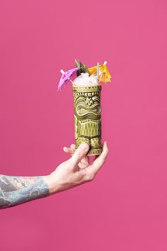 a person holding up a cup with flowers in it on a pink background that looks like tiki