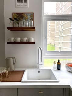 a white kitchen sink sitting under a window next to a wooden shelf filled with dishes