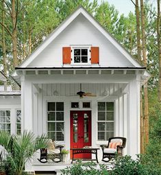 a small white house with red doors and shutters on the front porch is surrounded by greenery