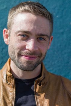 a close up of a person wearing a leather jacket and smiling at the camera with a blue wall in the background