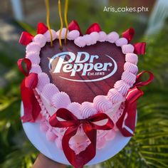 a heart shaped cake with dr pepper on top and red ribbon around the edges is held up by someone's hand