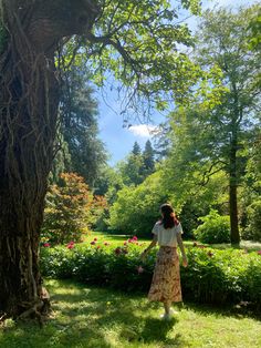 a woman walking through a lush green park next to a tree and shrubbery on a sunny day