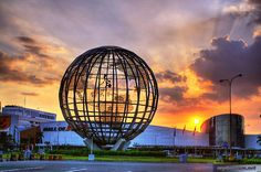 the sun is setting behind a large metal ball in an empty parking lot with buildings