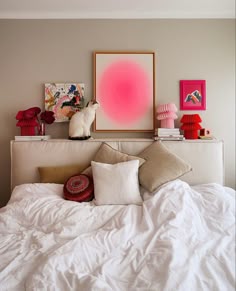 a white bed topped with lots of pillows next to a pink painting on the wall