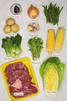an assortment of vegetables and meats on a white counter top, including corn, cabbage, green beans, broccoli, onion, carrots, celery, lettuce