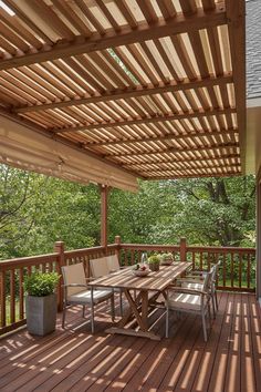 a wooden deck with table and chairs under a pergolated roof over looking a wooded area