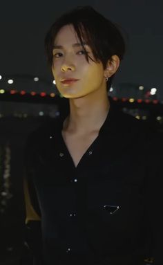 a young man standing in front of a body of water at night with the city lights behind him