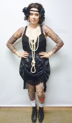 a woman with tattoos wearing a black dress and pearls on her head is standing in front of a white wall