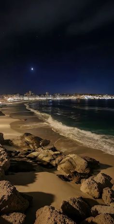 the beach at night with some rocks in the foreground and city lights in the background